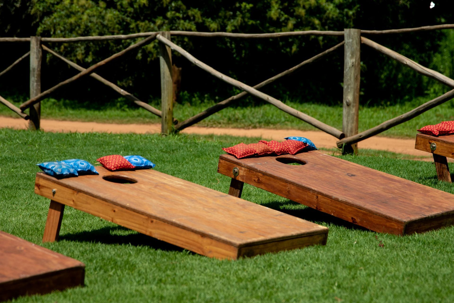 Wooden cornhole boards with blue and red bean bags sit on a grassy lawn in front of a rustic wooden fence.