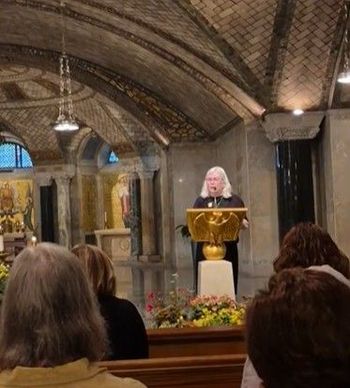 Speaker at a golden lectern in a vaulted stone chapel, viewed from the pews.