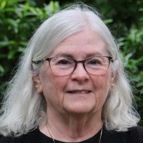Portrait of an older woman with white hair and glasses, smiling outdoors against greenery