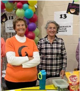 Two women at a table in front of balloons and numbered signs at a charity event