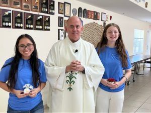Three people in blue shirts stand with a priest in a church hall, holding flowers and smiling.