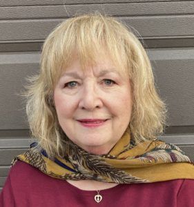 Smiling woman with blonde hair wearing a maroon top and patterned scarf against a gray wall