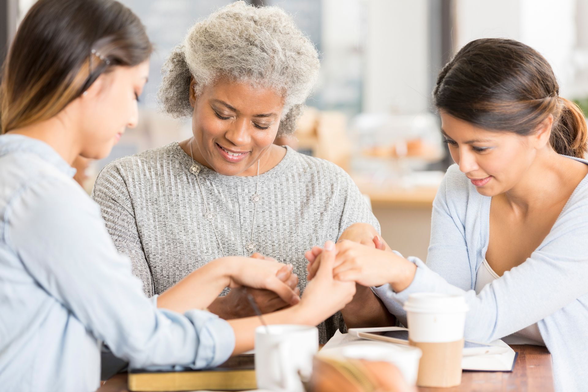 Three people sharing a warm, focused moment around a table, holding hands in a bright room