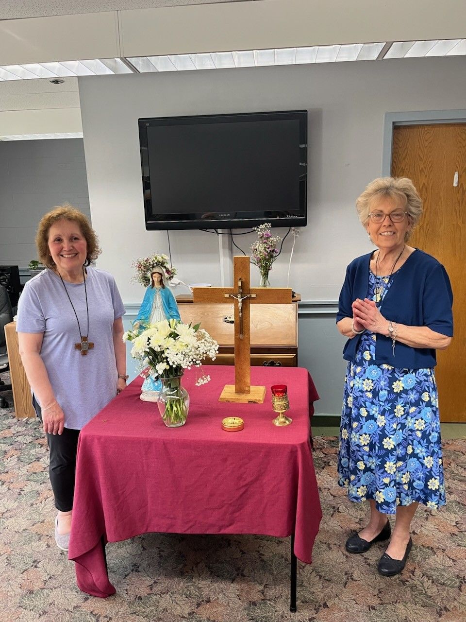 Two women stand beside a table with flowers, candles, and a cross in a room with a wall-mounted TV.