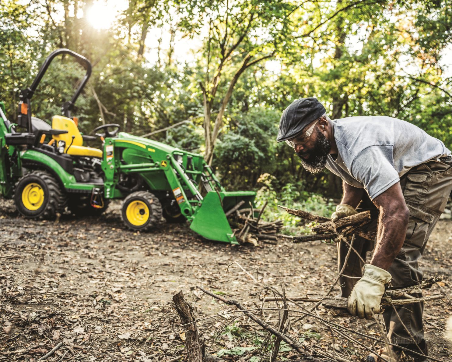 A man picks up limbs and loads them into the loader bucket of a 1025R John Deere compact tractor.