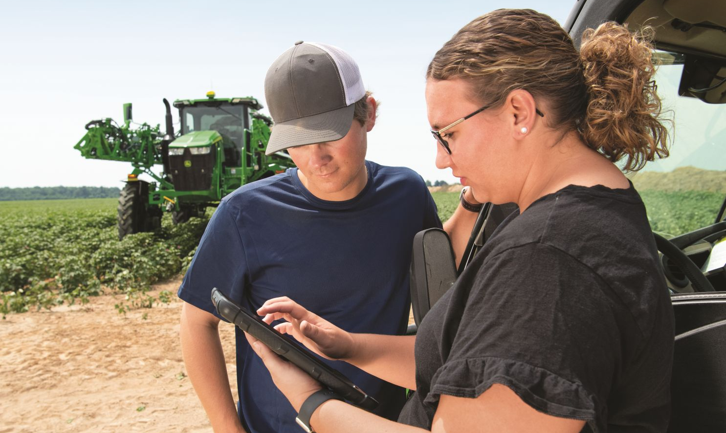 man and woman look at an iPad in the field with John Deere equipment nearby