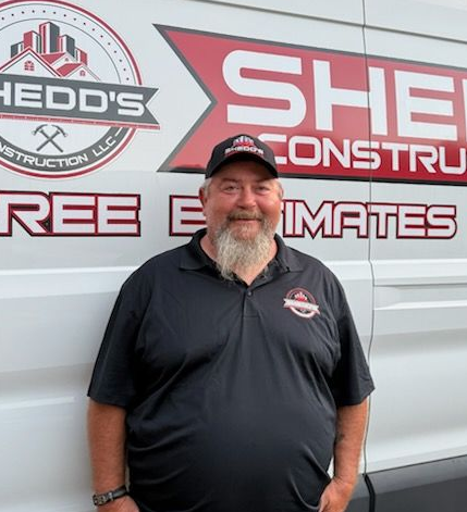 Smiling man in black shirt and cap standing in front of a Shedd Construction truck