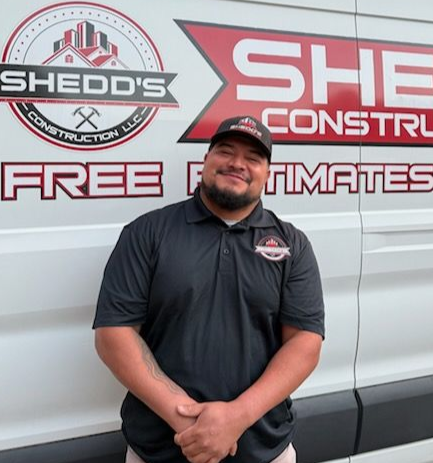 Man in black polo and cap standing in front of a Shedd’s Construction van with “Free Estimates” signage