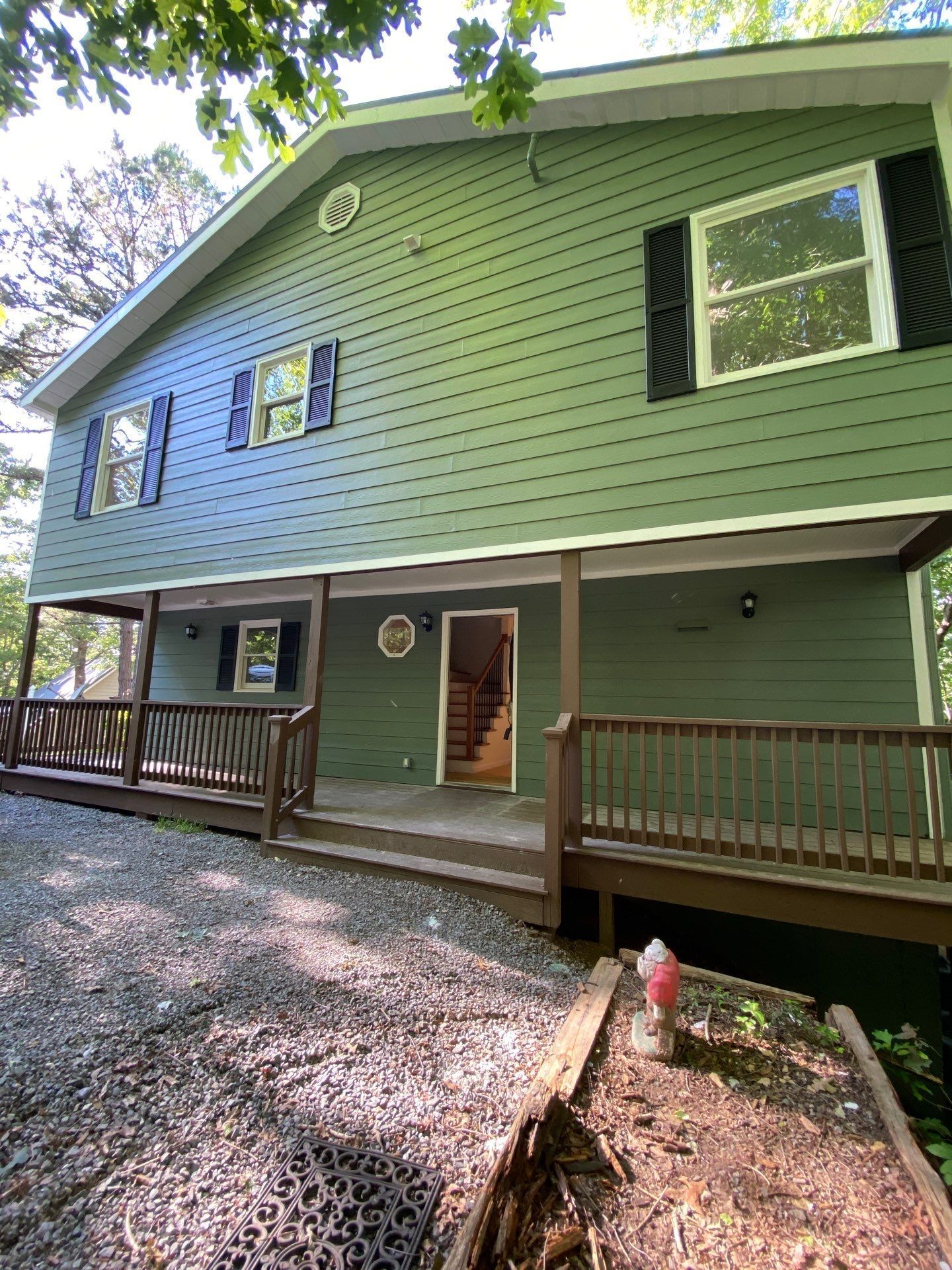 Green two-story house with a wooden porch and railing, viewed from a gravel driveway under trees