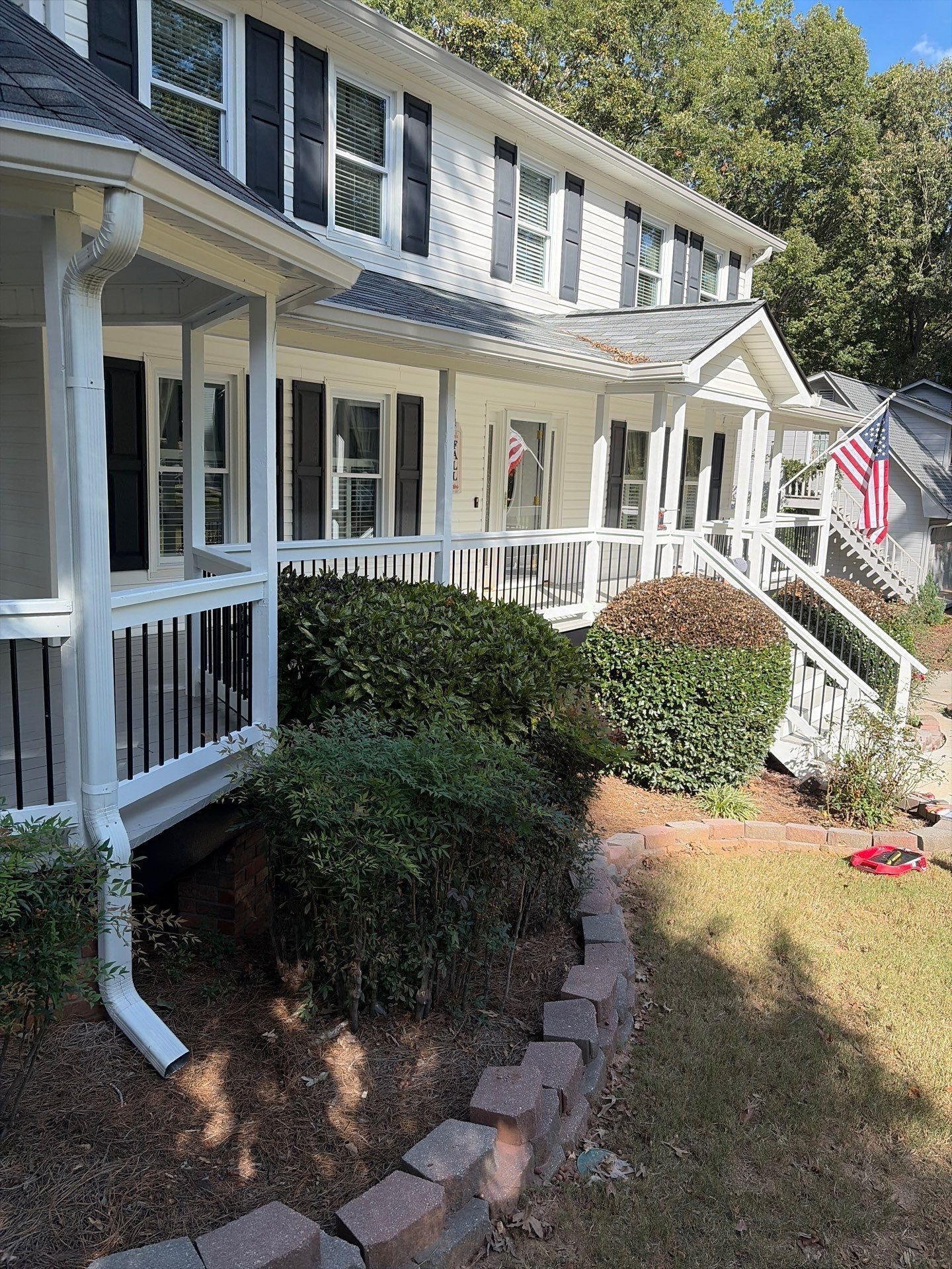 Two-story house with white porch railings, shrubs, stone edging, and an American flag by the front steps.