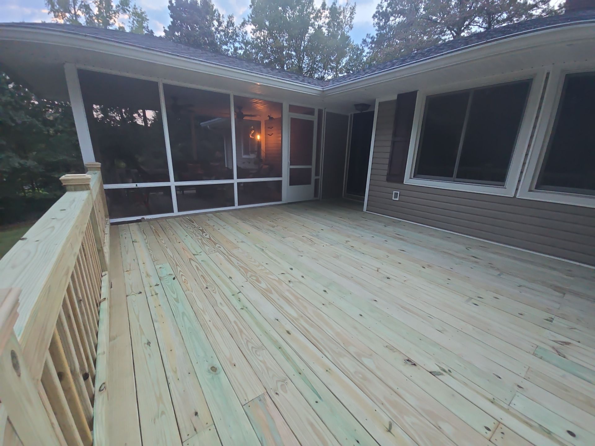 Spacious wooden deck with white railings beside a house, looking toward a screened porch at dusk