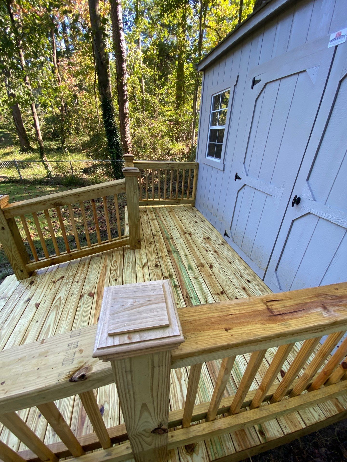 Wooden deck with railing beside a light blue shed in a wooded backyard.