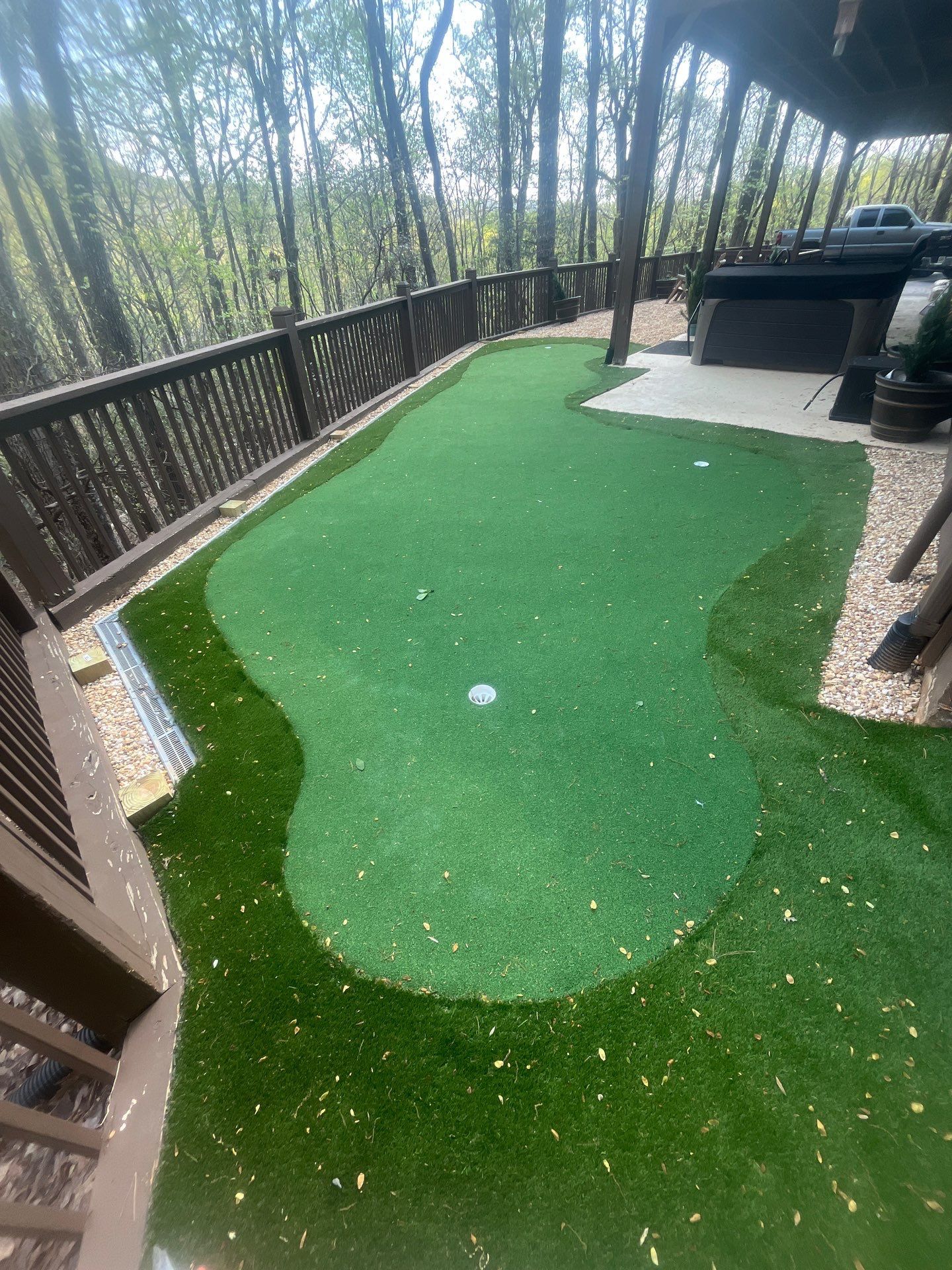 Backyard mini-golf green with a putting hole, wooden railing, and grill on a deck near trees