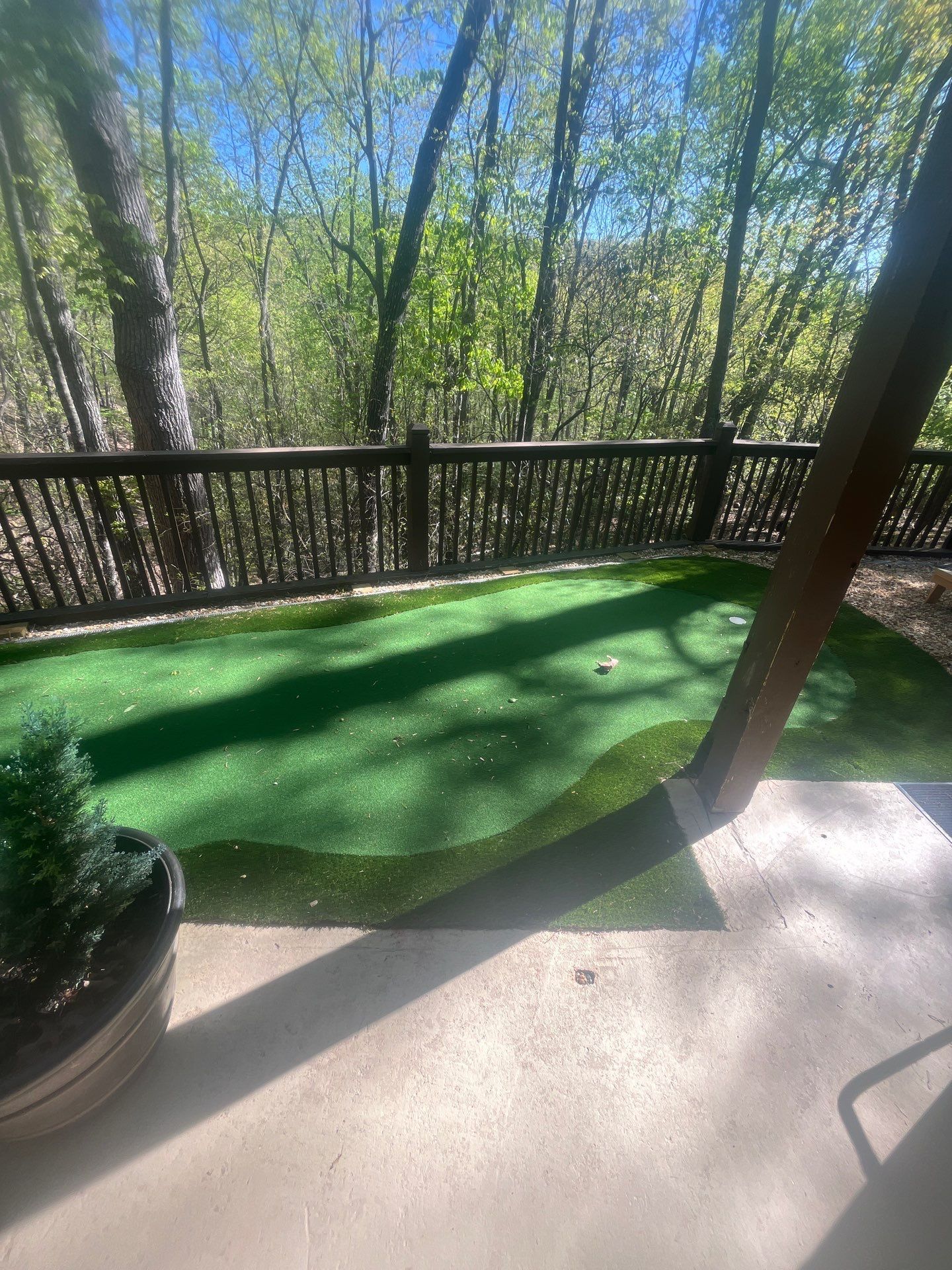 Wooden deck with a railing overlooking trees and a green artificial turf area in sunlight.