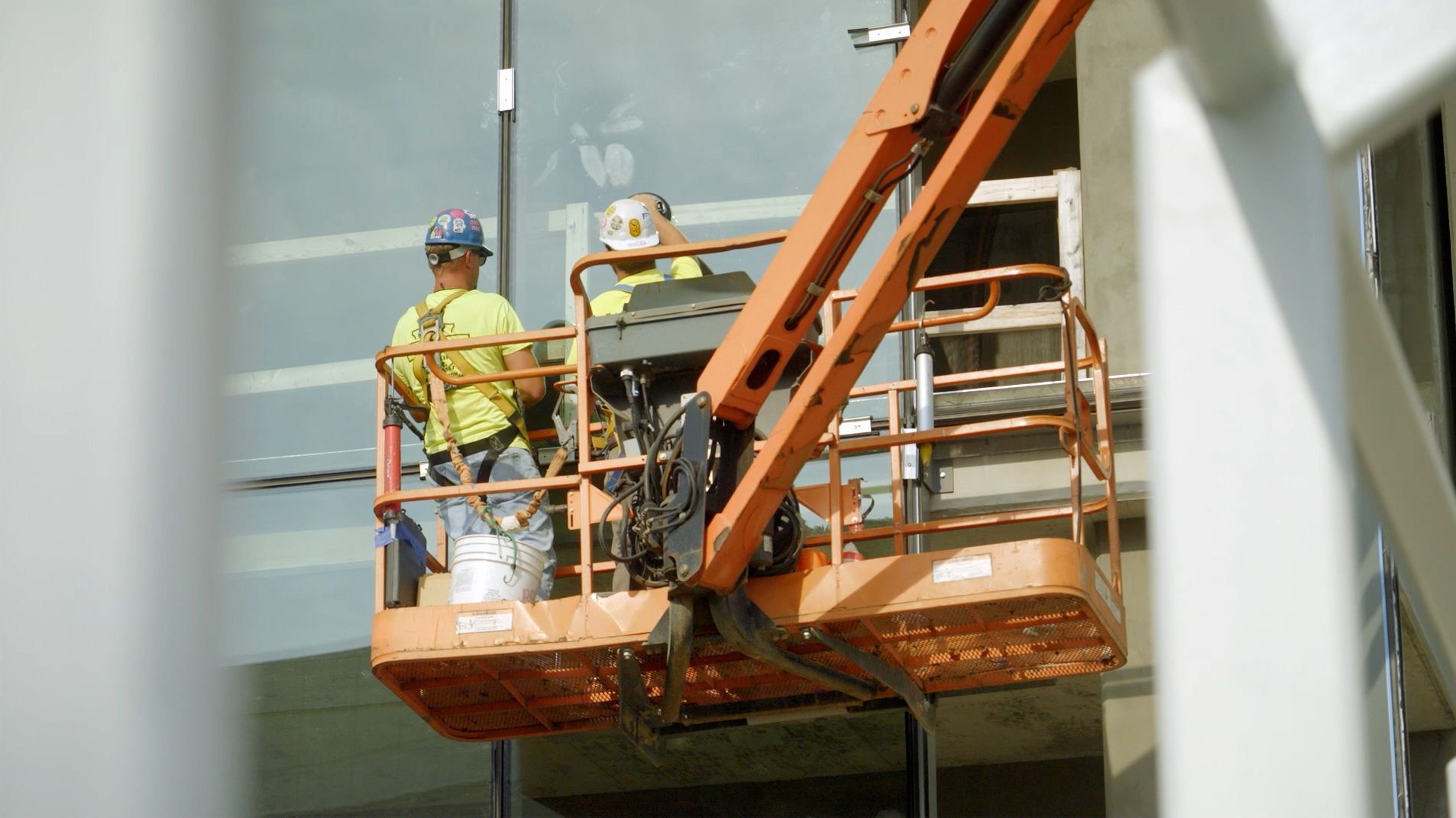 Glaziers operating an orange ariel lift to install large-scale glass panels on a building exterior.