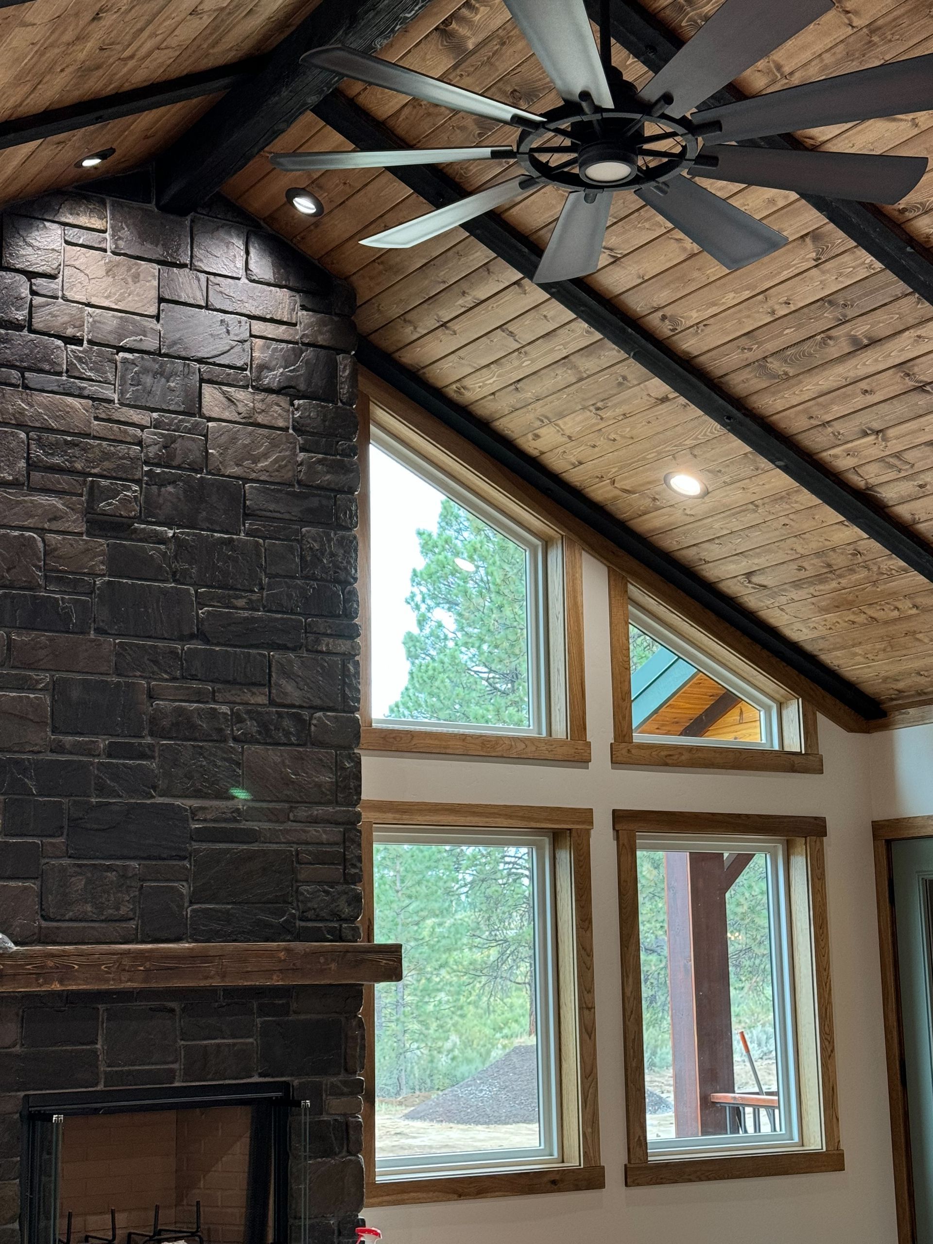 Rustic living room with stone fireplace, tall windows, vaulted wood ceiling, and ceiling fan.