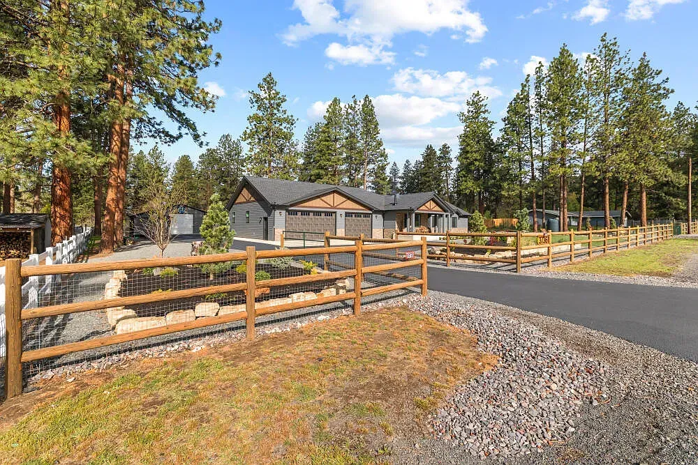 Wooden ranch gate and gravel driveway leading to a cabin among pine trees under a blue sky.