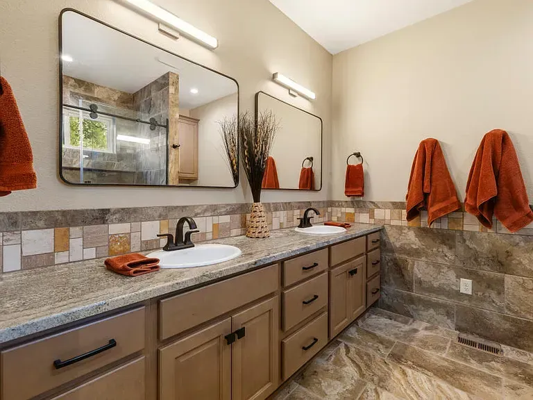 Modern bathroom with double vanity, large mirror, stone tile, and orange towels