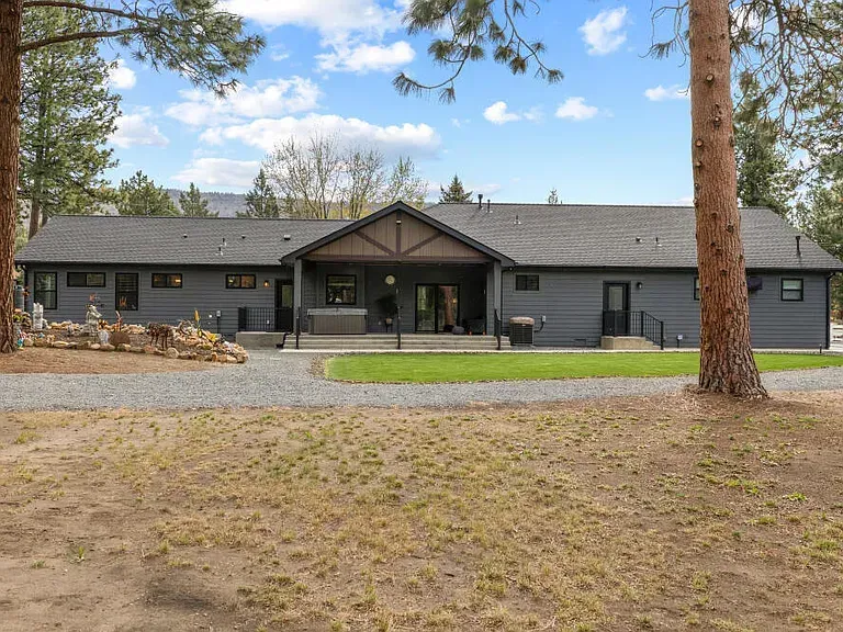 Gray ranch-style house with a covered front porch, gravel driveway, and tall trees in front