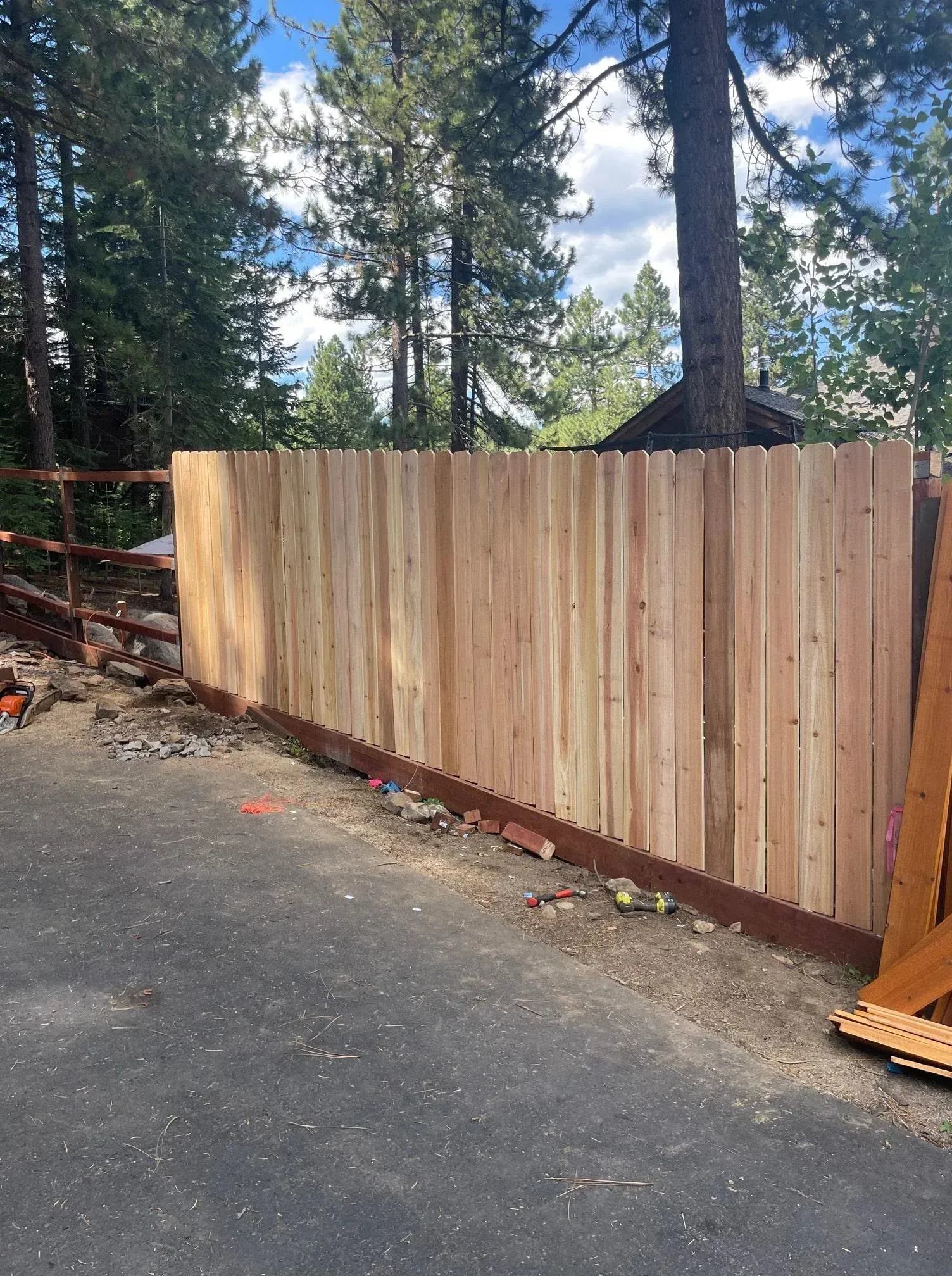 Wooden fence along a paved driveway with dirt and debris, with trees in the background.