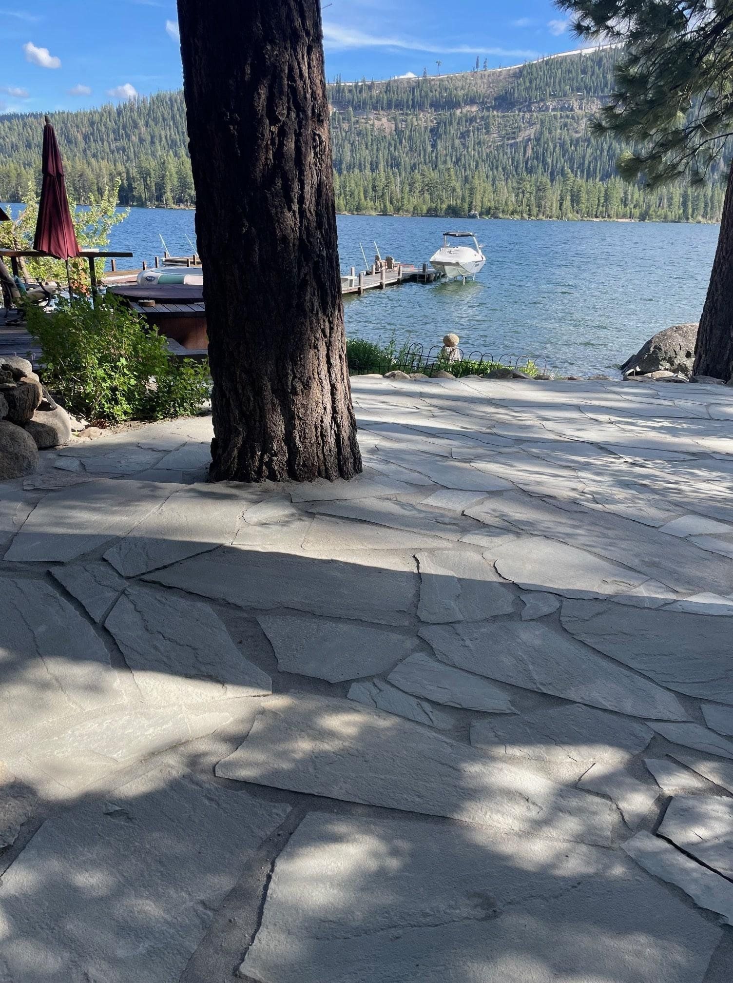 Stone patio leads to a lake with a boat, trees and mountains in the background. Sunny day.