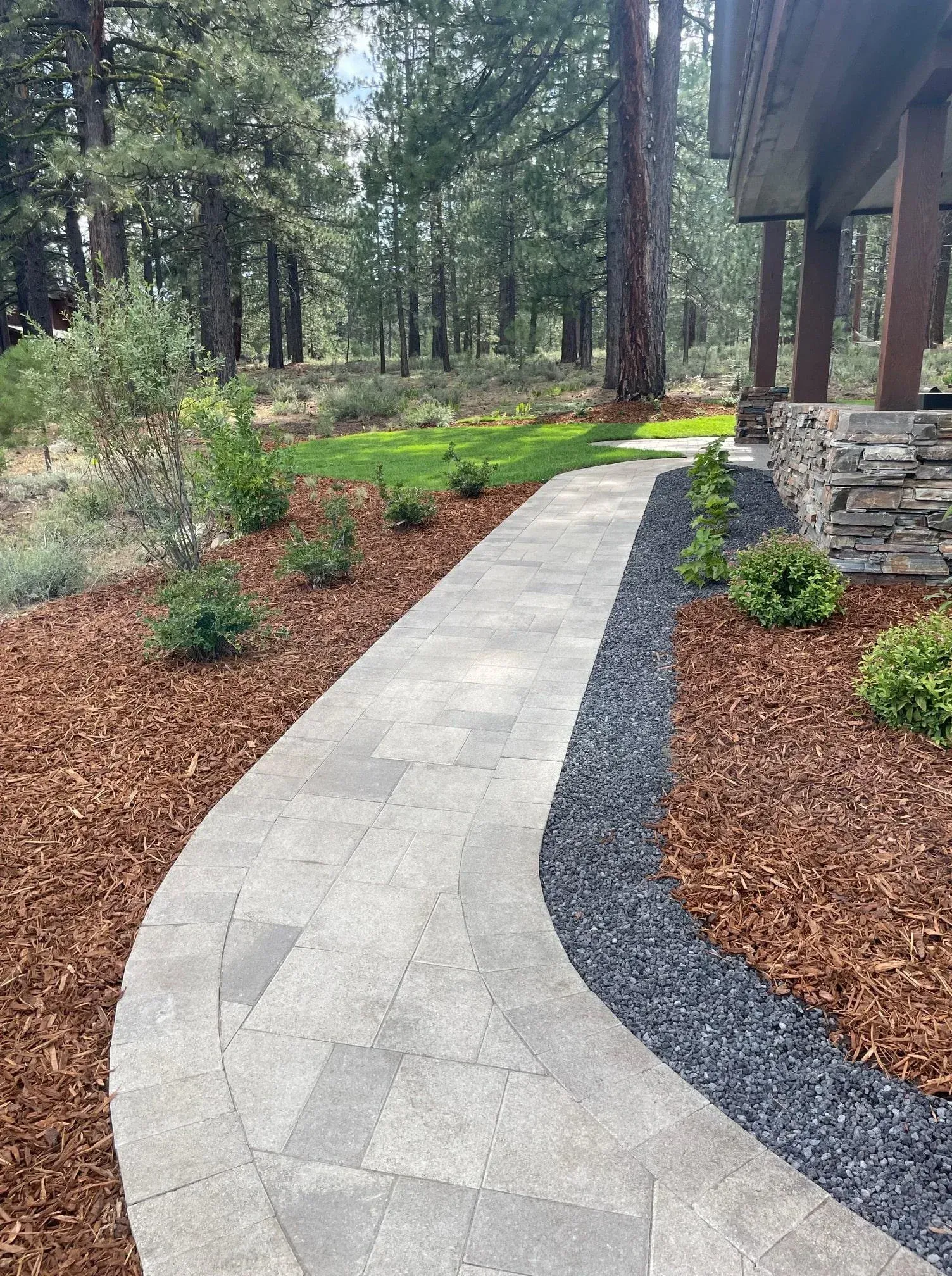 Stone walkway curves through a landscaped yard with mulch, plants, and black rock border. Forest in background.
