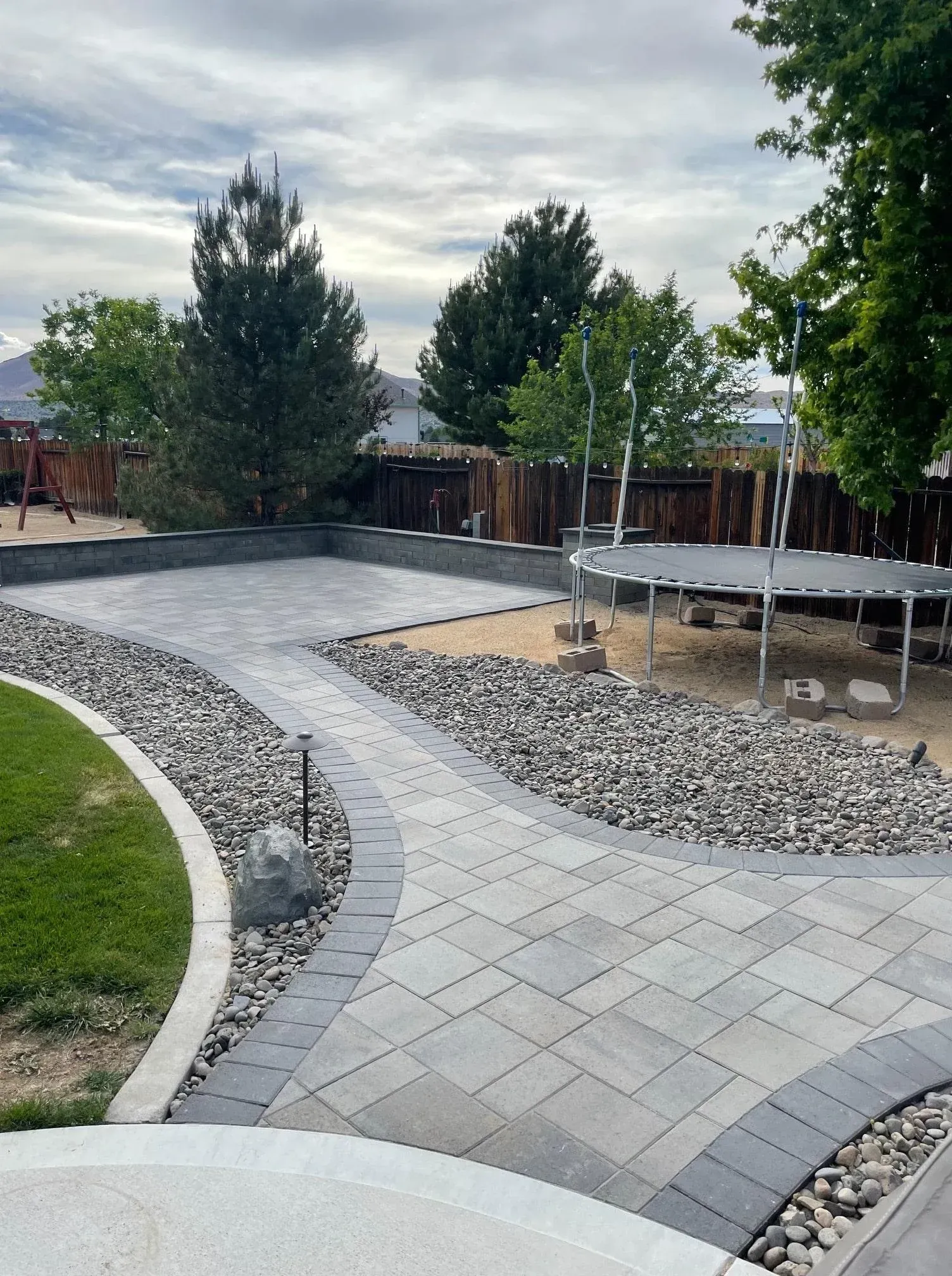 Backyard with paved path, rock beds, trampoline, and trees under cloudy sky.