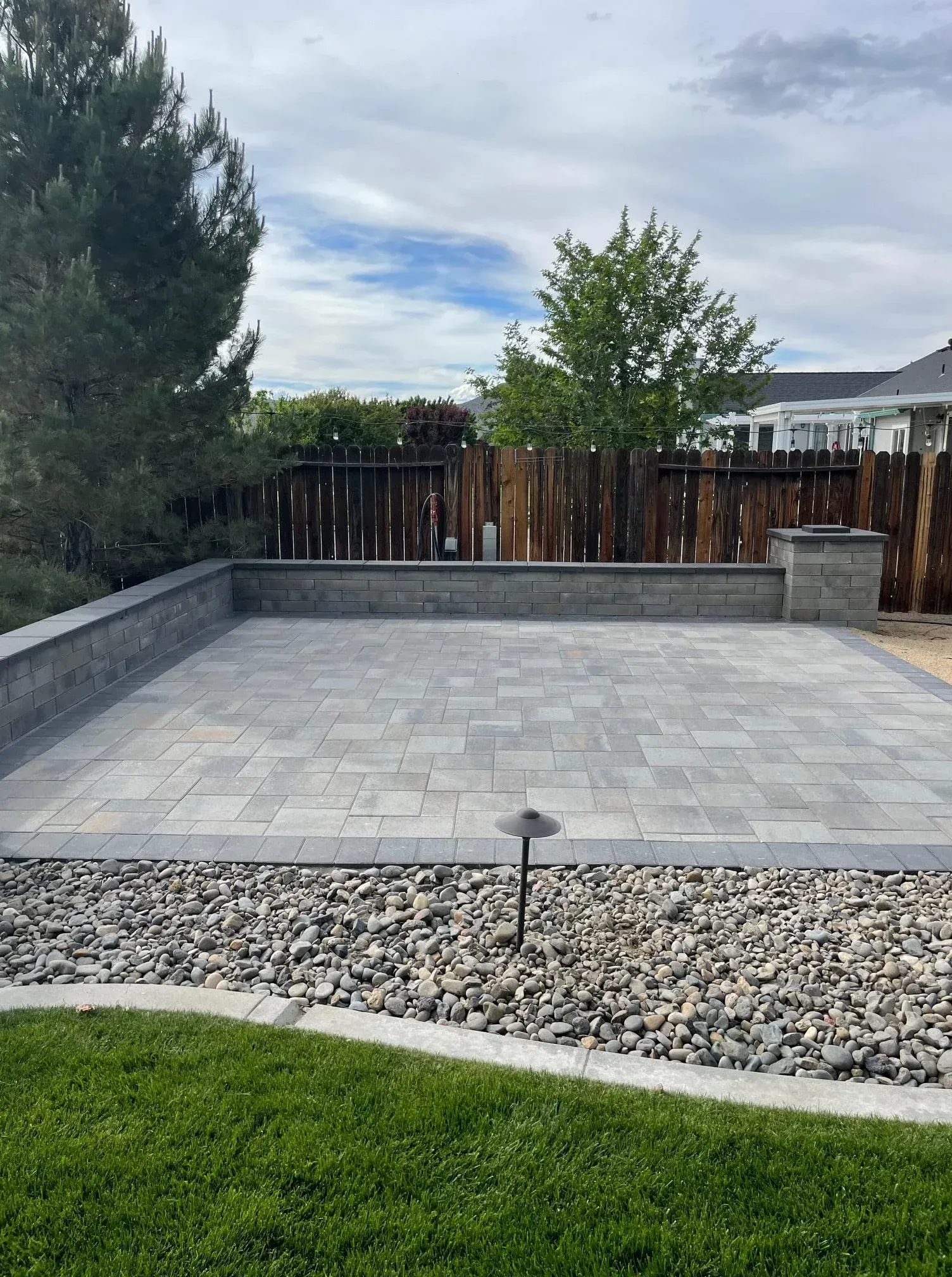 Patio paved with gray tiles, surrounded by a low wall, gravel, and grass. Cloudy sky.
