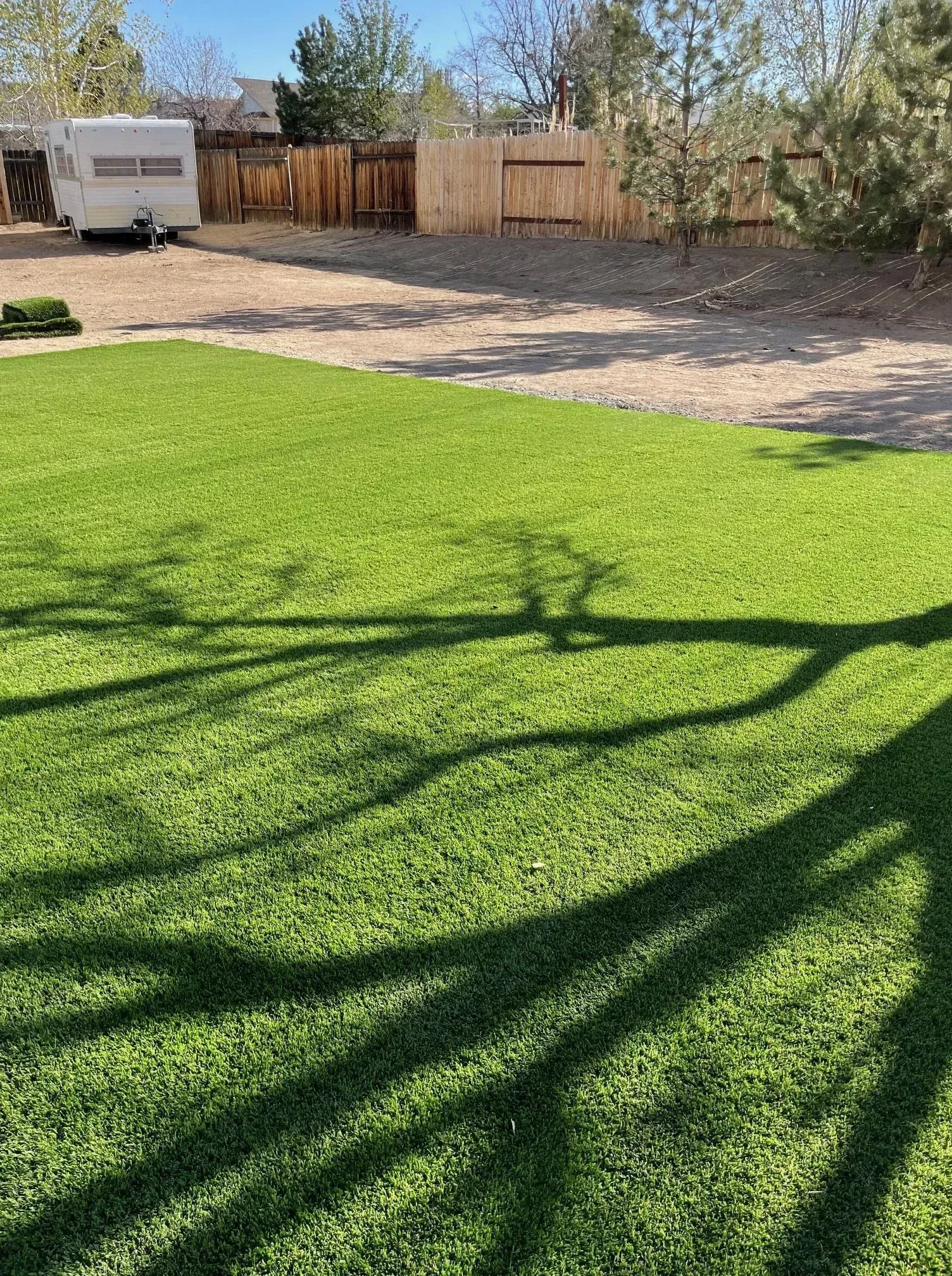 Green lawn with tree shadows, gravel driveway, and a camper next to a wooden fence.