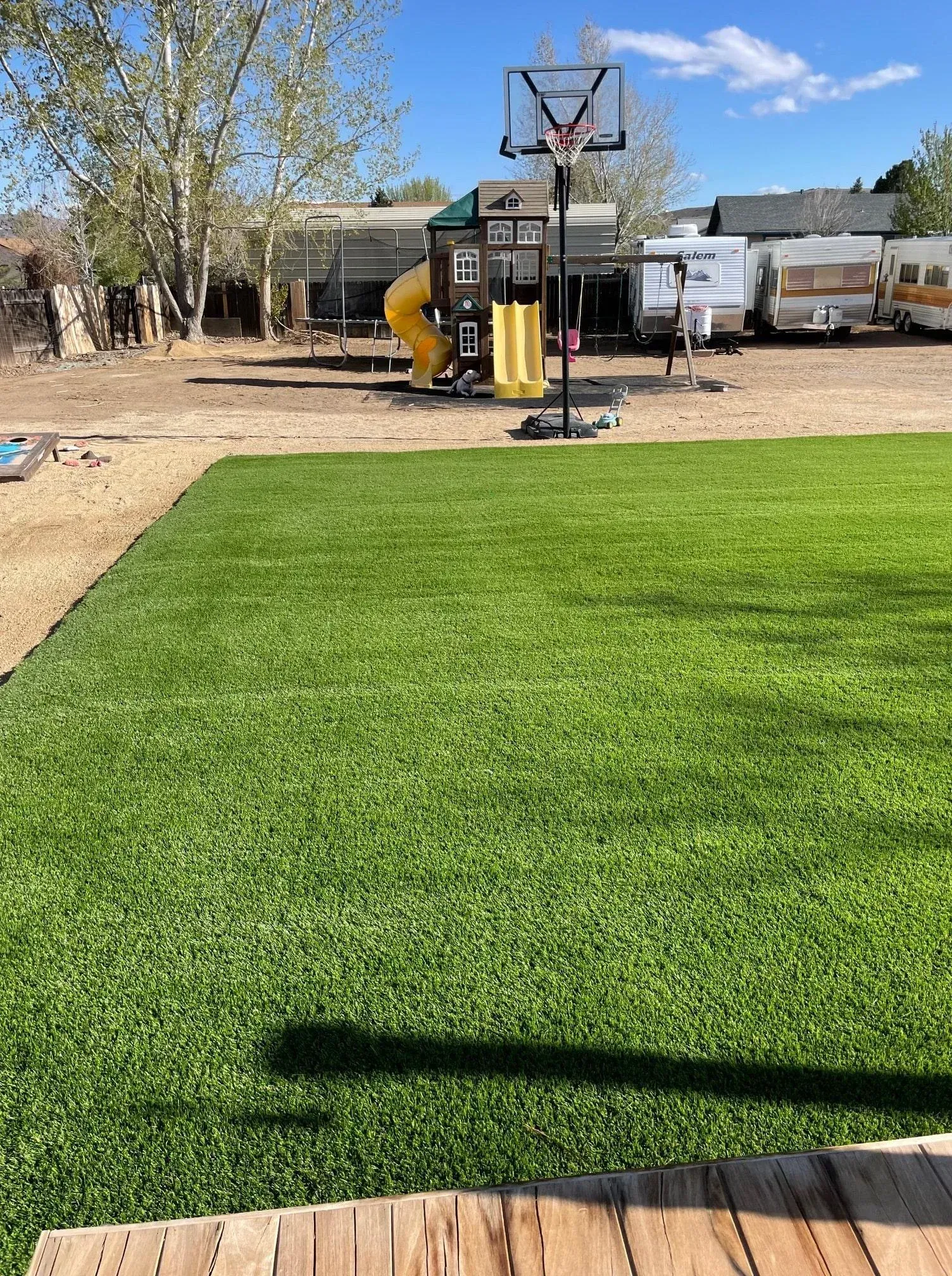 Green artificial grass with a playground, basketball hoop, and RVs in the background under a blue sky.