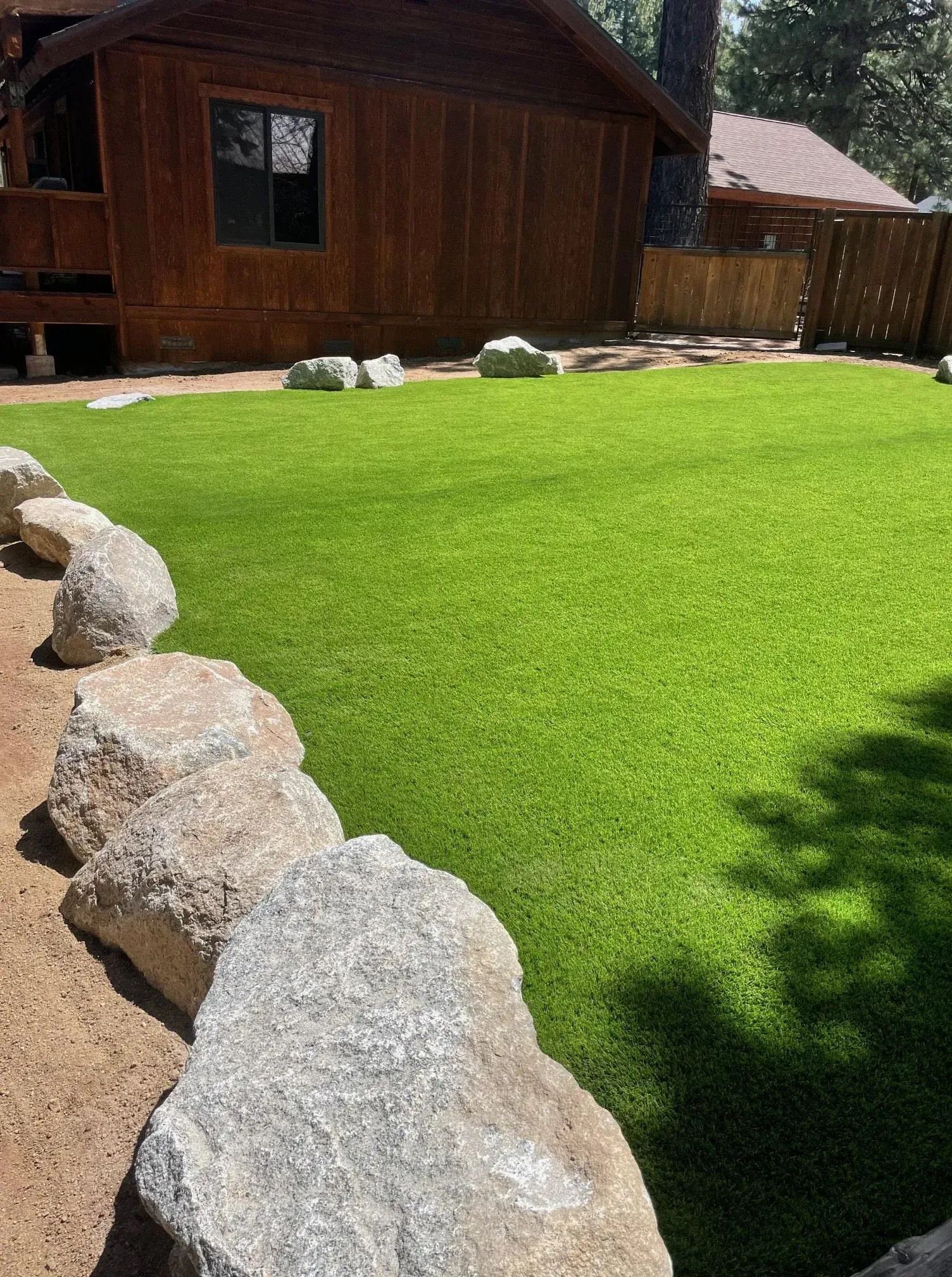 A vibrant green artificial lawn bordered by large rocks, in front of a wooden cabin.