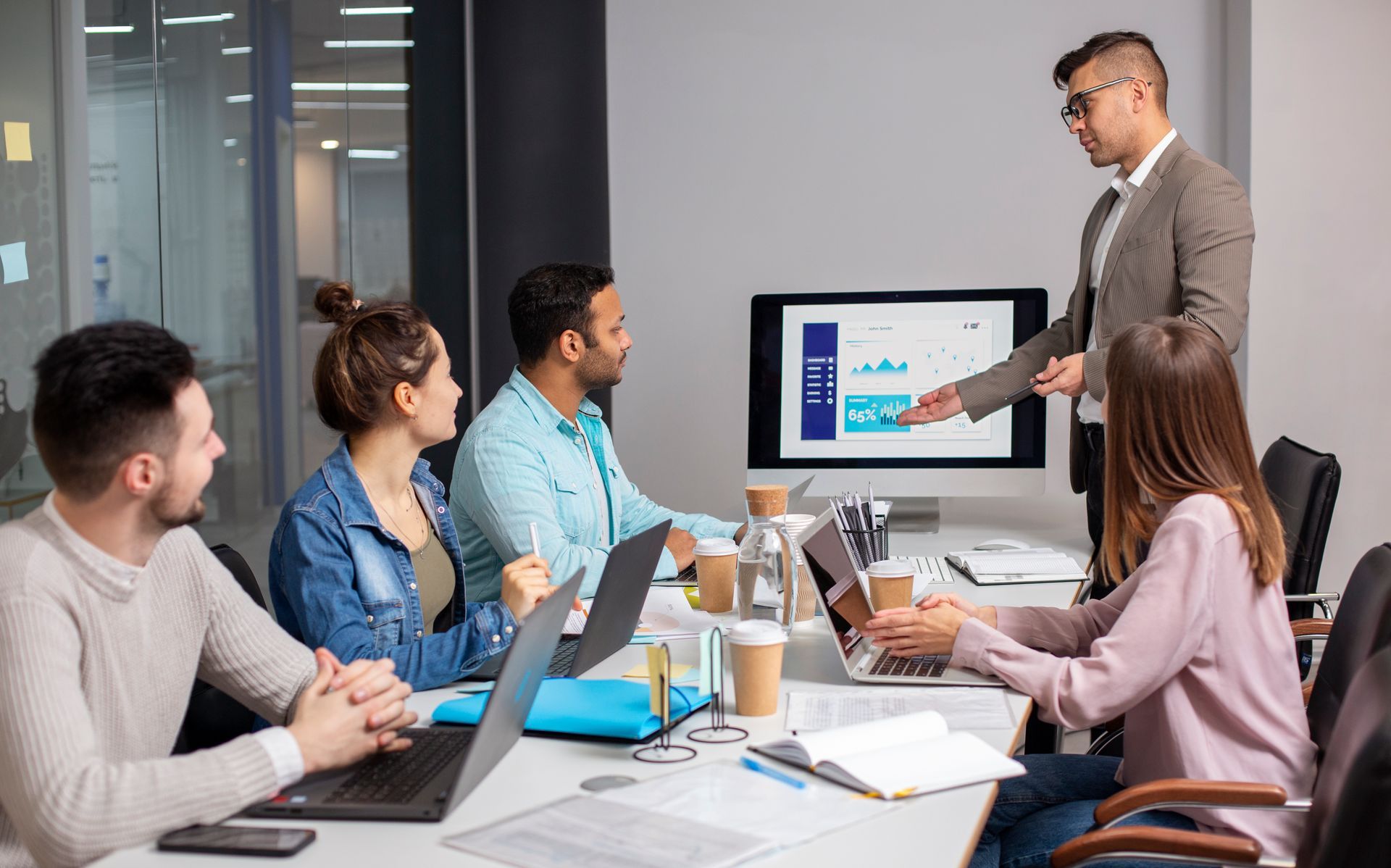 Group of people in an office meeting, man presenting data on a monitor, others looking on.