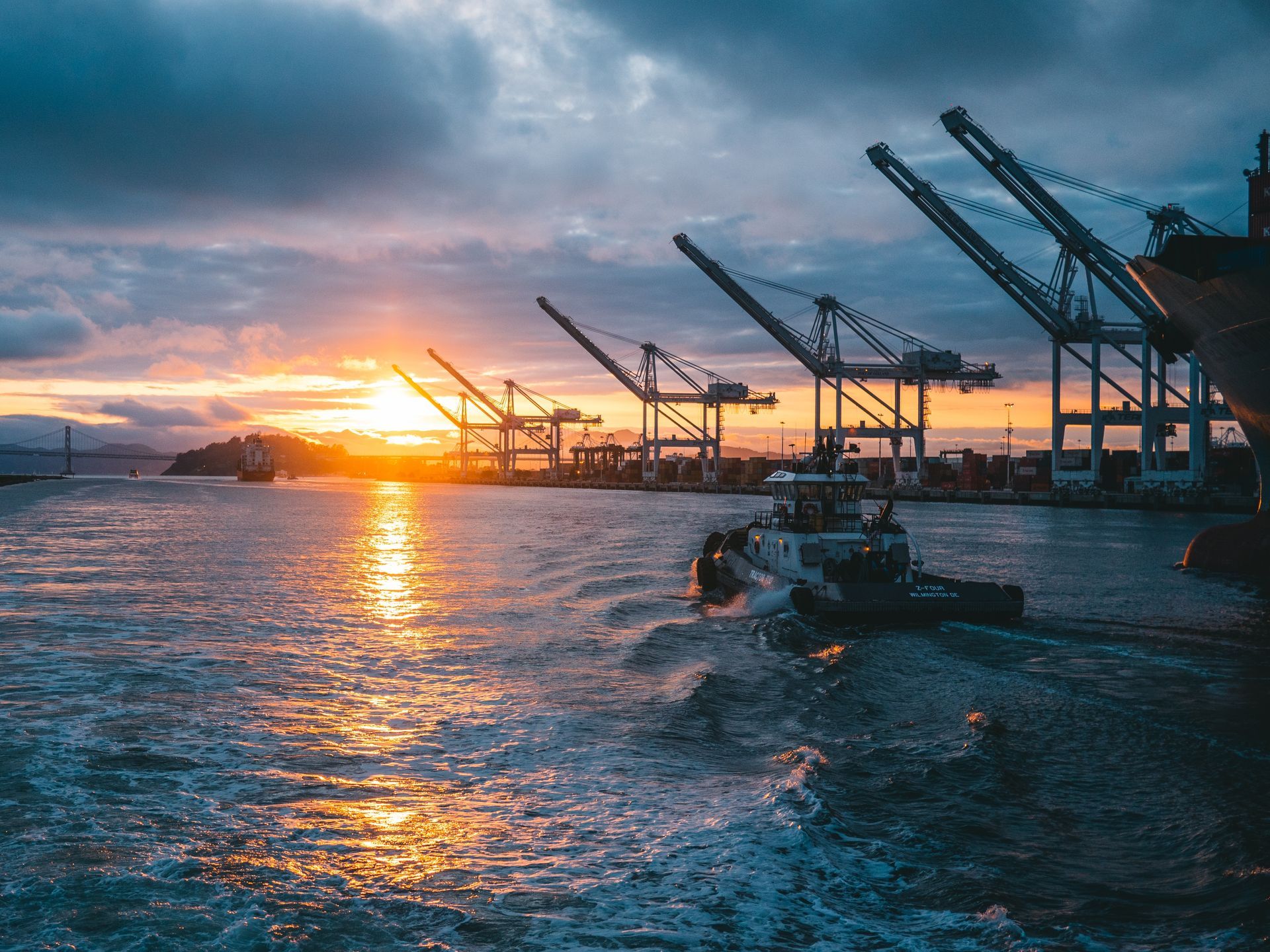 Tugboat departing a container port at sunset with ship to shore cranes and stacked cargo containers along the quay