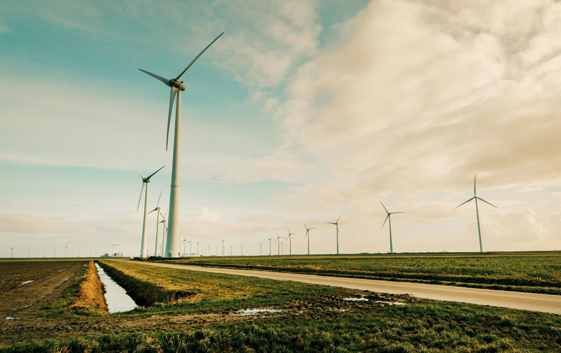 Wind turbines in a field under a cloudy blue sky with a dirt road and irrigation ditch.