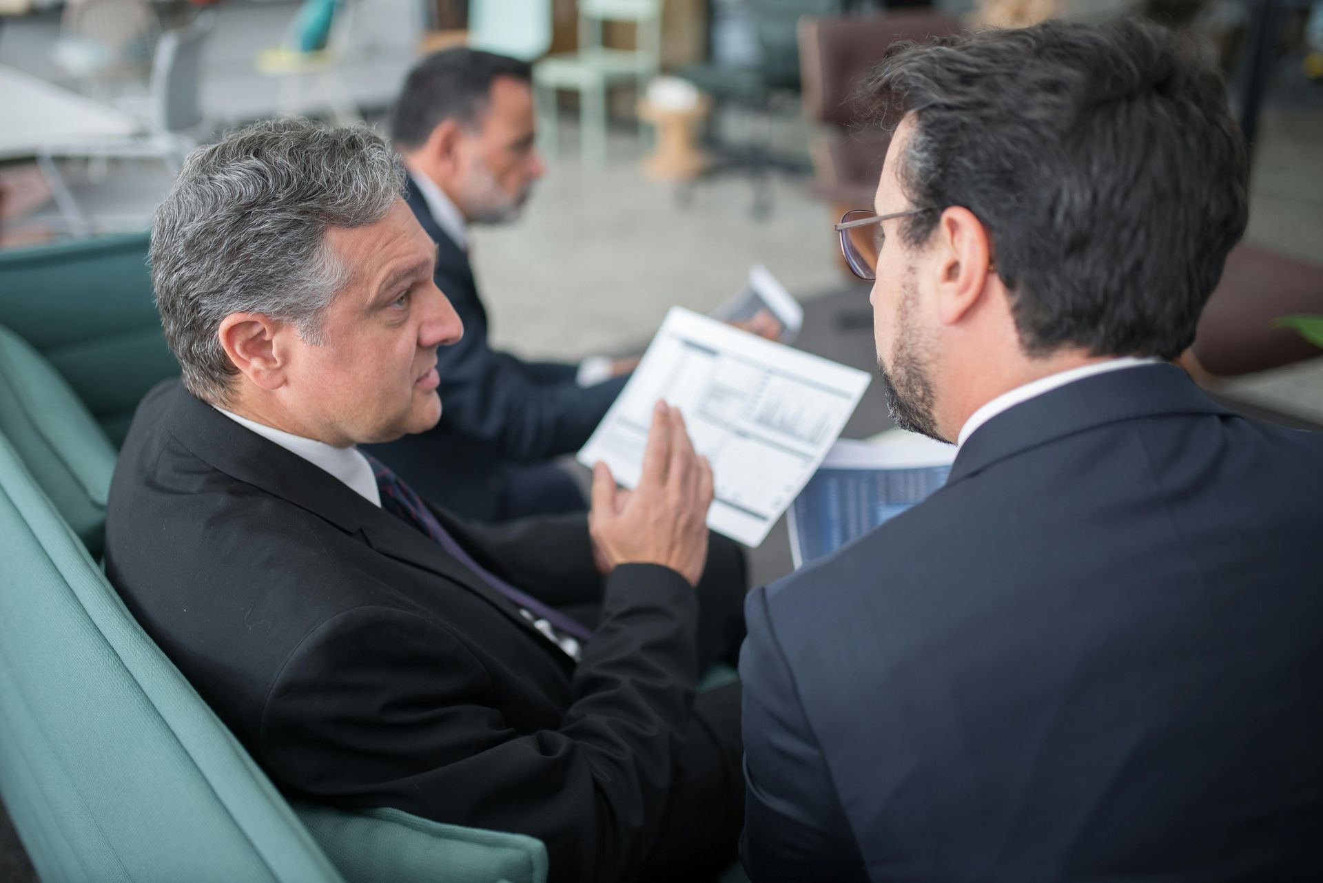Three businessmen in suits reviewing papers, discussing business in an office lounge.