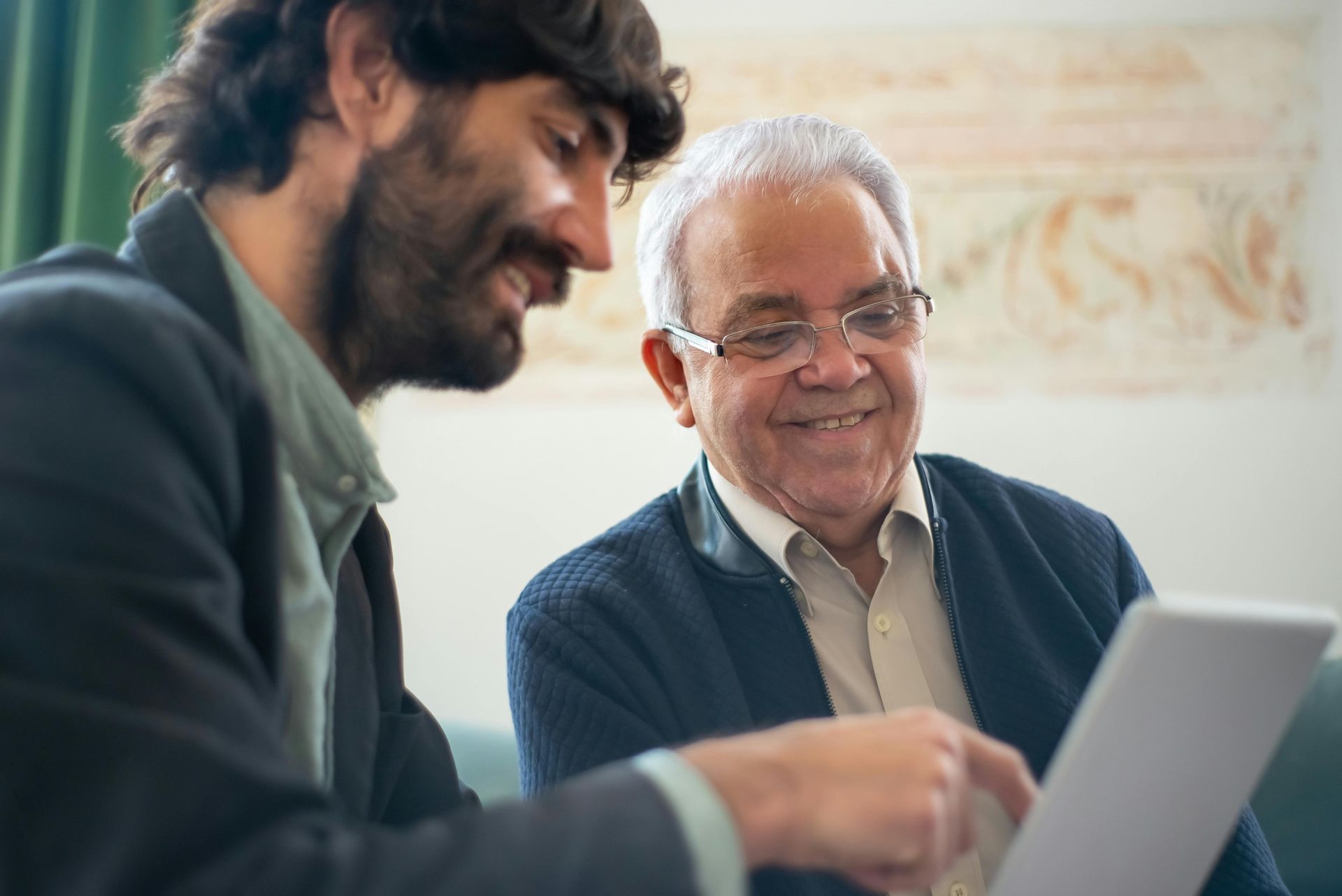 Young man showing elderly man a tablet, both smiling.