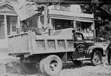 Black and white photo of a dump truck being loaded with a large container of material near a house.