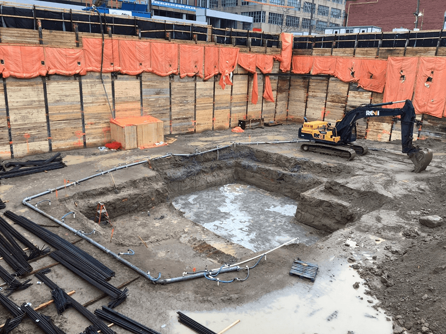 Chantier de construction avec une petite excavatrice dans une fosse rectangulaire; des barrières en bois et orange entourent.