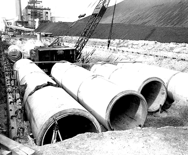 Concrete pipes in trench at construction site, with crane and building in background.