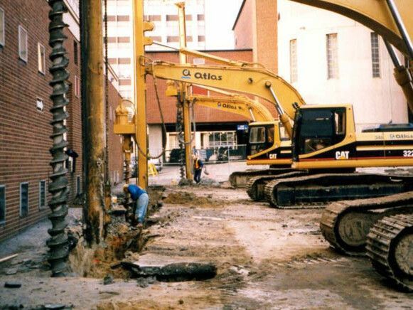 Chantier de construction avec des excavatrices jaunes et des ouvriers qui forent près d'un bâtiment.