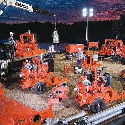 Workers setting up orange water pumps at an outdoor worksite at dusk. A crane is lifting equipment.