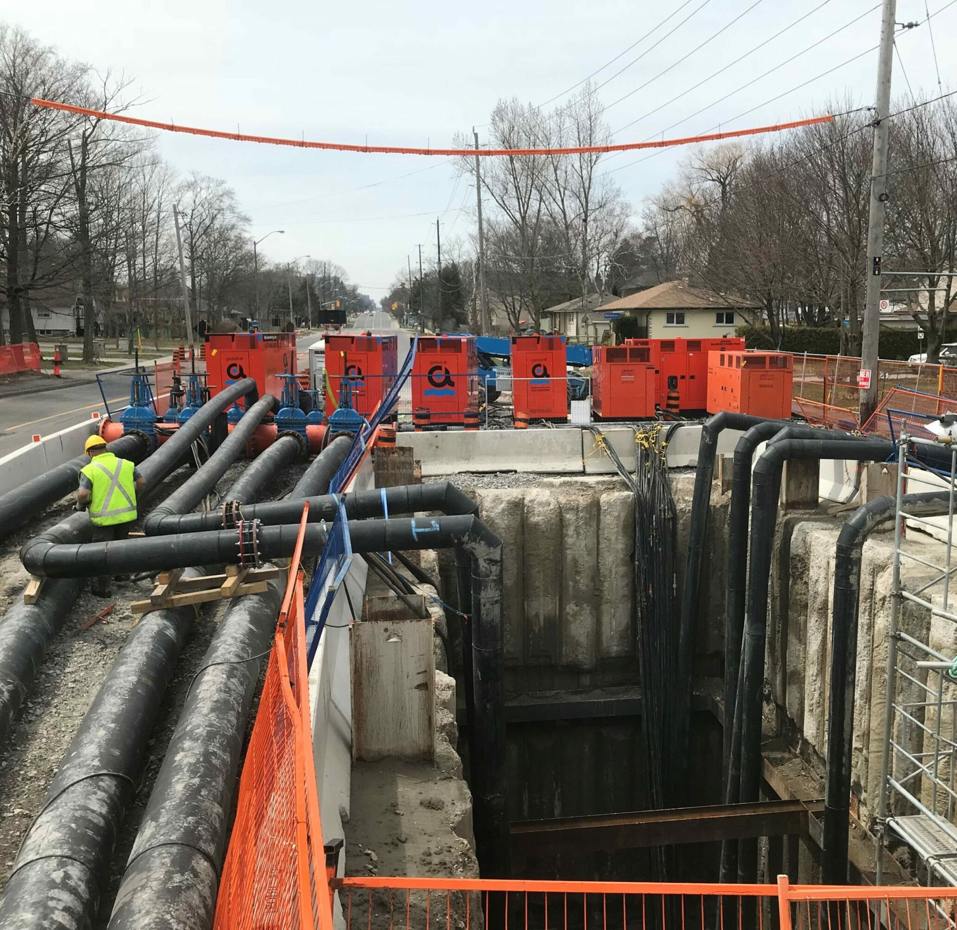 Chantier de construction avec des ouvriers, des machines orange et des tuyaux noirs posés sur une route.