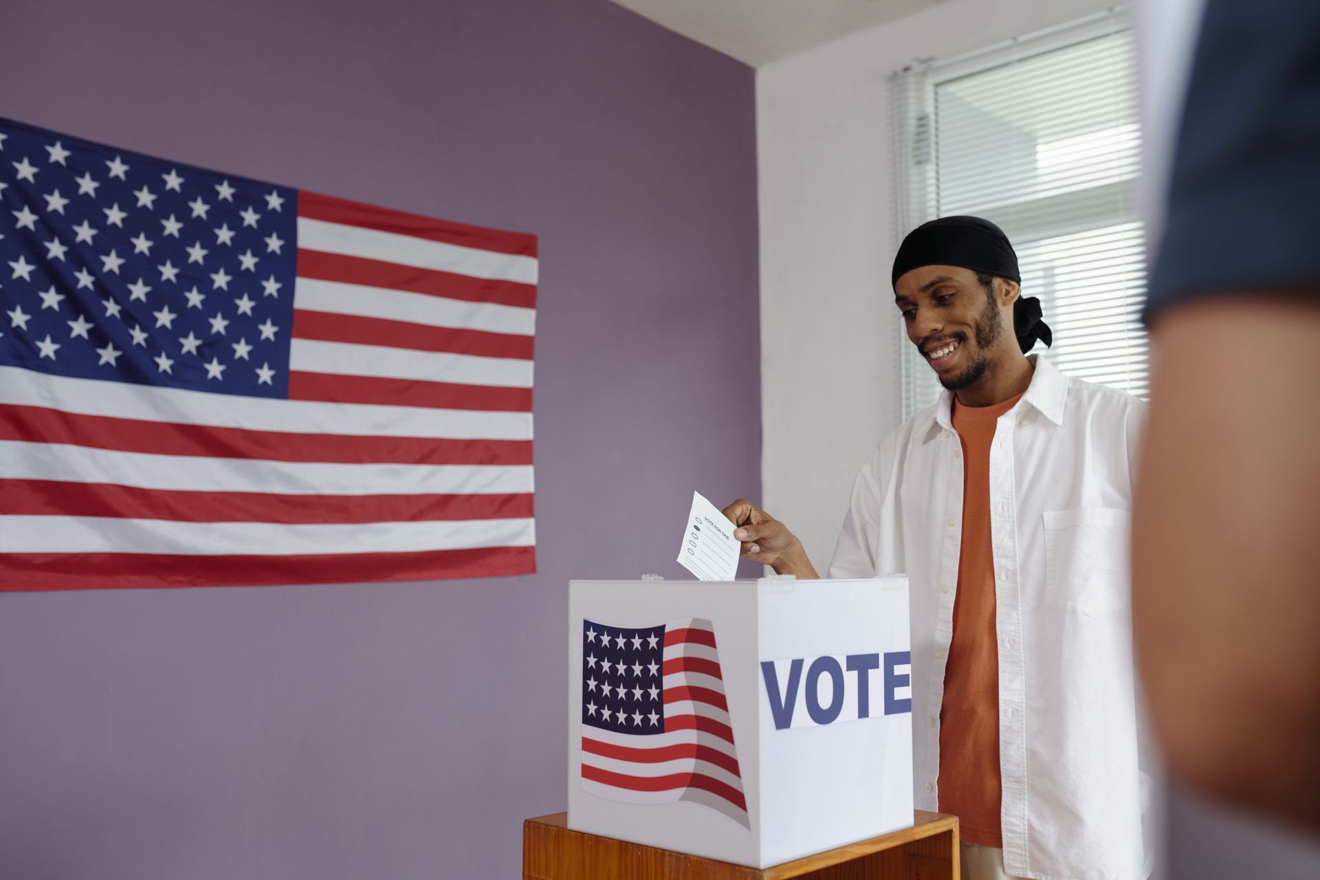 Person standing beside a voting booth with an American flag on the wall, smiling and holding a ballot