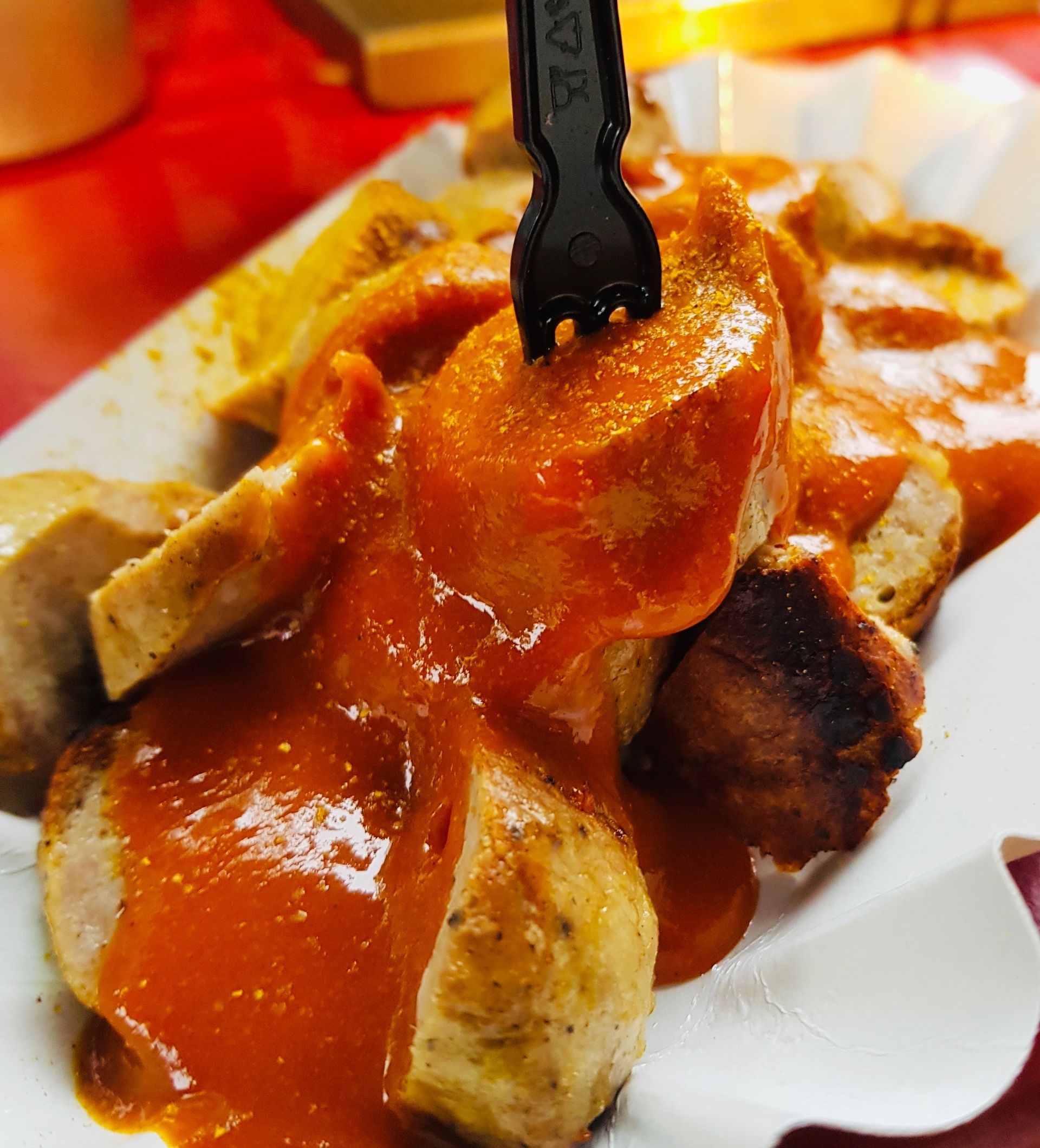 Nahaufnahme von Bratwurst mit rotem Curry-Ketchup auf einem Papiertablett, durchstochen von einer schwarzen Plastikgabel.
