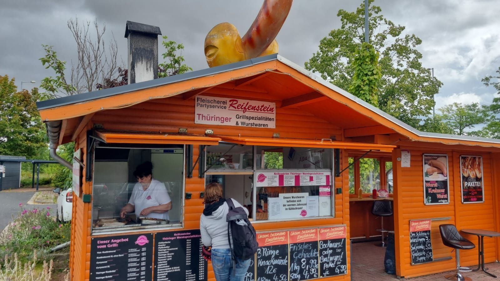 Ein kleiner Wurststand aus Holz mit einer großen Wurstskulptur auf dem Dach bedient einen Kunden an einem Kiosk im Freien.