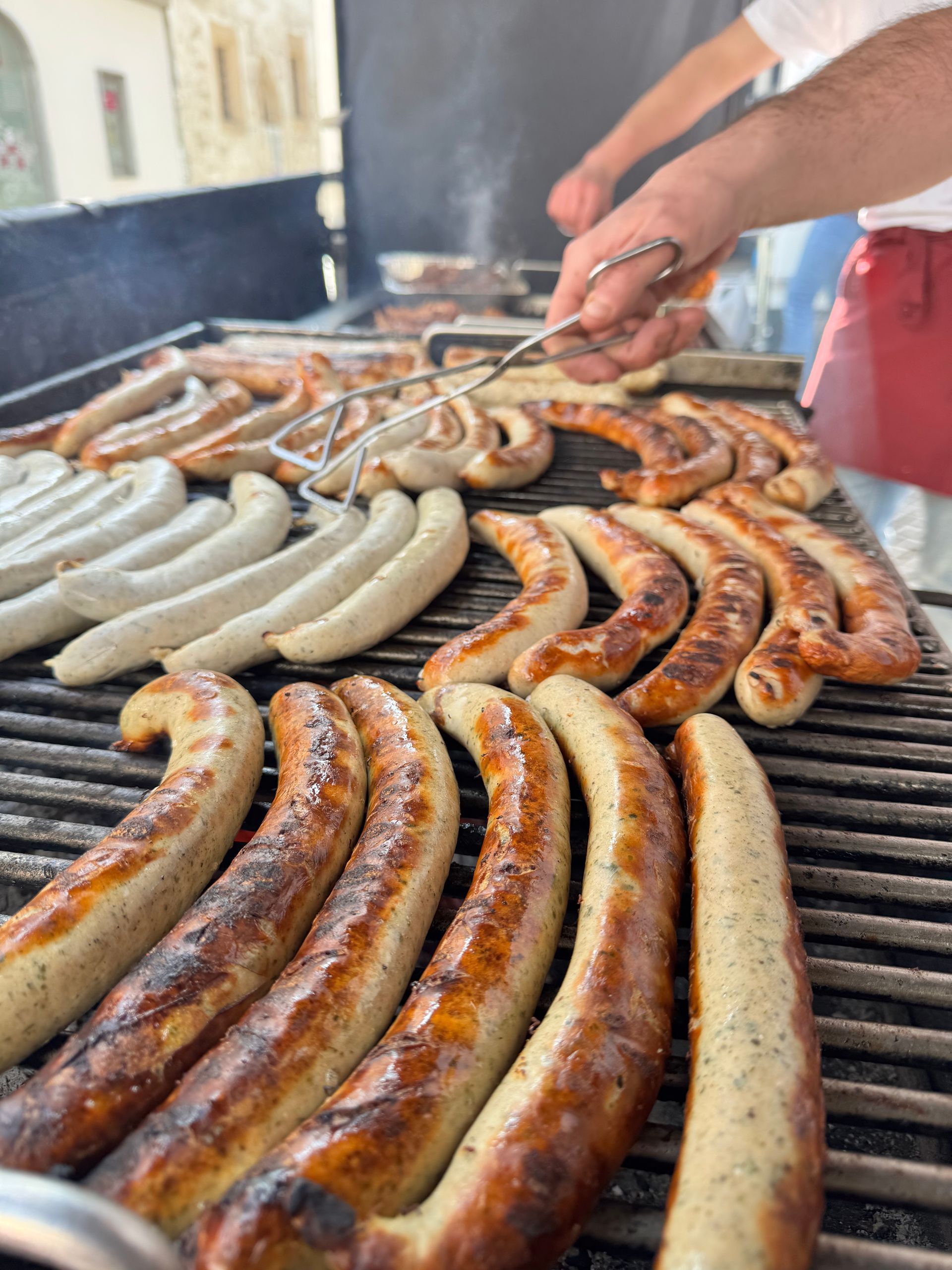 Eine Person wendet mit einer Metallzange Reihen von bräunenden Würstchen auf einem großen Außengrill.