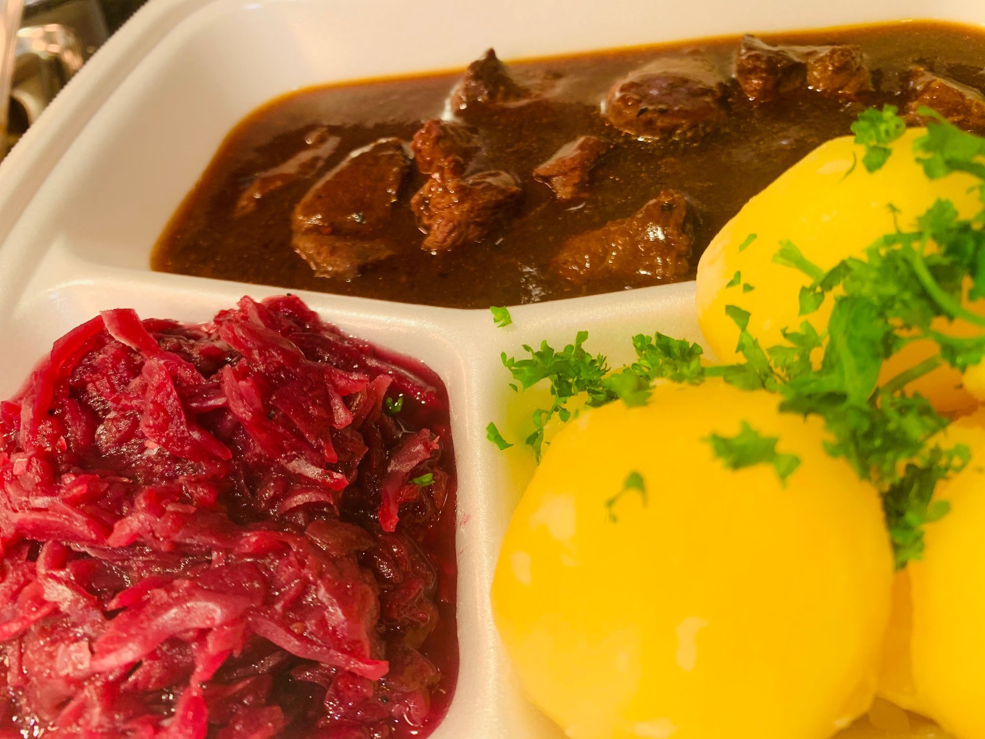 Eine Schale mit mit Rindfleischeintopf, Rotkohl und zwei gelben Kartoffelknödeln, garniert mit frischer Petersilie.