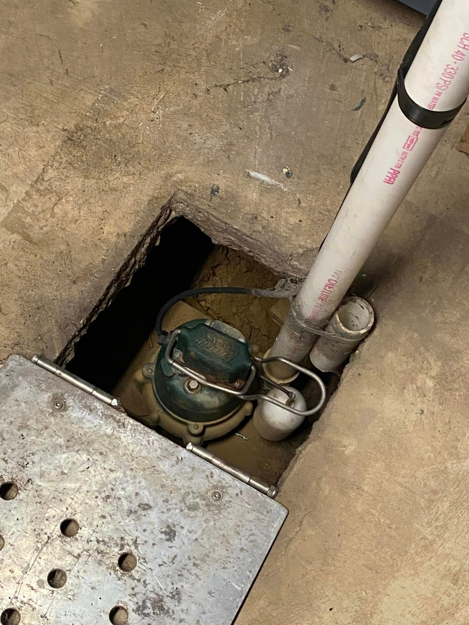 A sump pump is sitting in a hole in the floor next to a pipe.