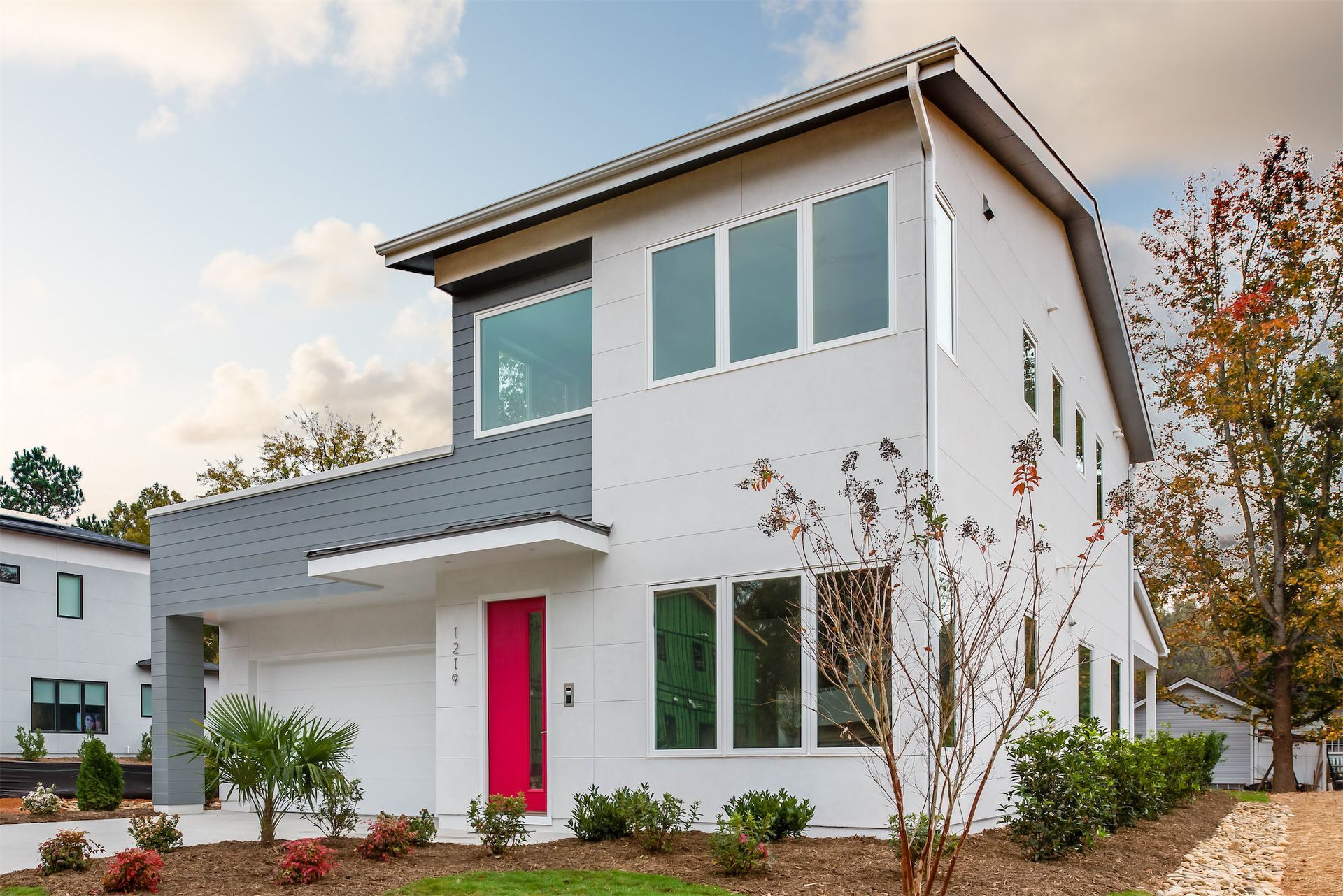 Modern two-story white house with a pink front door and large windows, surrounded by landscaping
