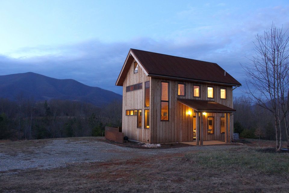 Small wooden house at dusk with warm lights, set in a rural field with mountains in the background